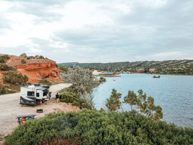 A white fifth-wheel camper is parked at a lakeside campsite with red rock cliffs and green shrubs under a cloudy sky.