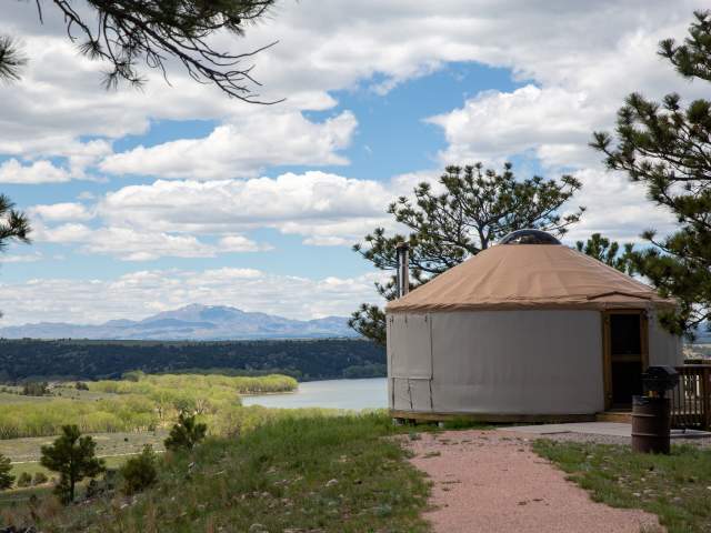 A circular tan and grey yurt sits on a grassy hilltop overlooking a scenic valley in Guernsey State Park, Wyoming.
