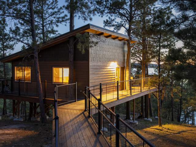 A modern wooden cabin treehouse with glowing windows sits nestled among pine trees, featuring an elevated deck and a long boardwalk entrance during the warm light of dusk.