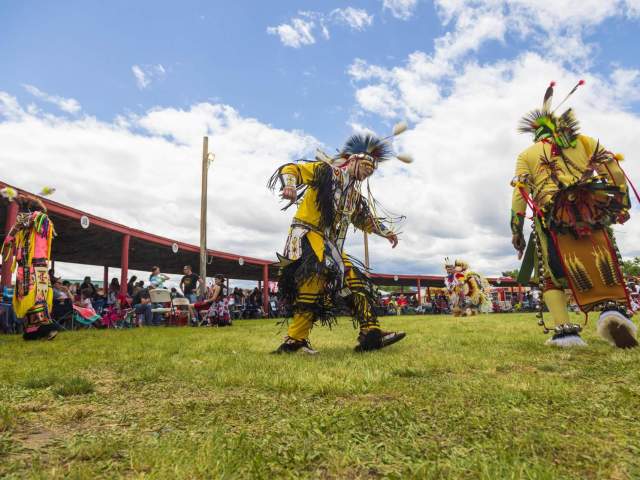 A group of men wearing traditional clothing and performing in a field surrounded by a crowd at the Annual Eastern Shoshone Indian Days Powwow.