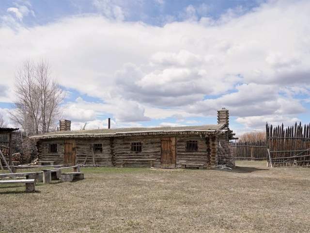 Historic homes sit in a grassy field, with a flag waving in the distance above the mountain ridge.