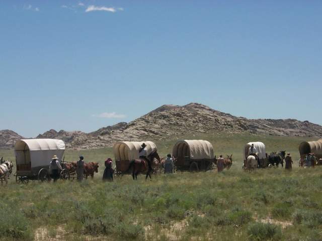 People stand beside covered wagons and horses in tall grass, with rugged mountains rising in the distance.