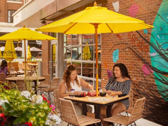 two women eating at an outdoor table at a restaurant sitting under a yellow umbrella with a mural on the wall behind them