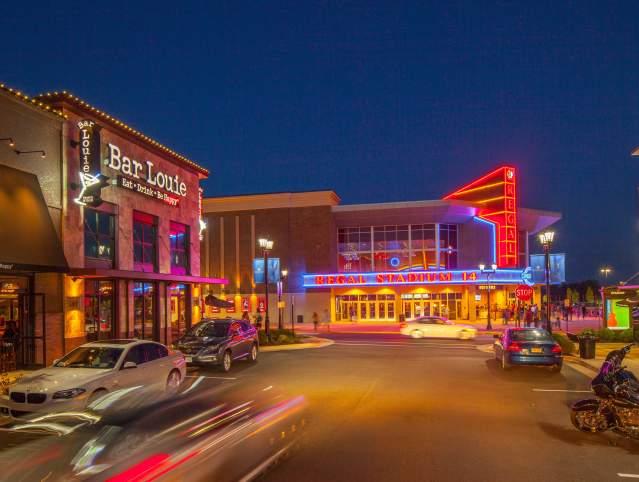 a shopping area with neon lights at night with cars driving by