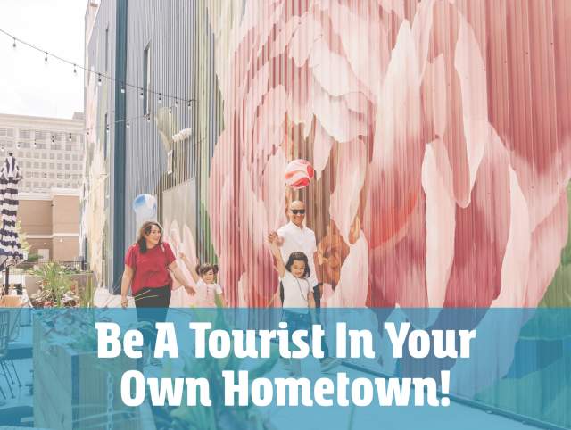 A family strolls along The Landing enjoys public art. Superimposed on the image is the title card for Be A Tourist In Your Own Hometown, the annual tourism discovery event in Fort Wayne, Indiana.