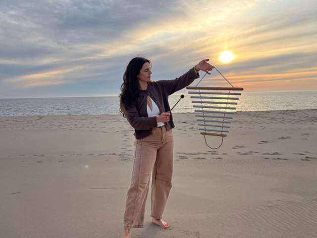 Person standing barefoot on a sandy beach during sunset, holding and playing a handheld wind chime instrument with a mallet. The ocean stretches across the background under a cloudy sky with warm orange light near the horizon. The person wears a jacket and light-colored pants, and faces slightly toward the instrument with arms raised, focusing on the chimes.