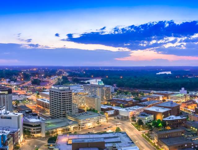 Panoramic sunset view of downtown Montgomery skyline with city lights, riverfront buildings, and Alabama horizon at dusk glow.