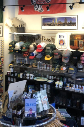 Interior of a Milwaukee-themed retail shop filled with local sports and city merchandise. Shelves and wall displays are packed with baseball caps, glassware, mugs, magnets, and souvenirs featuring teams like the Bucks and Brewers. Framed posters, vintage signs, and banners line the upper walls, while a central display table holds books and additional items. The space is brightly lit and densely arranged, creating a colorful, curated storefront focused on Milwaukee pride.