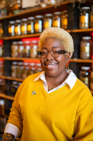Angela Mallett is smiling, standing behind the counter of HoneyBee Sage. She's wearing a bright yellow sweater over a white collared shirt, along with gold jewelry including earrings and a necklace. Behind her are wooden shelves lined with numerous glass jars containing various spices, herbs, or other ingredients with labels.