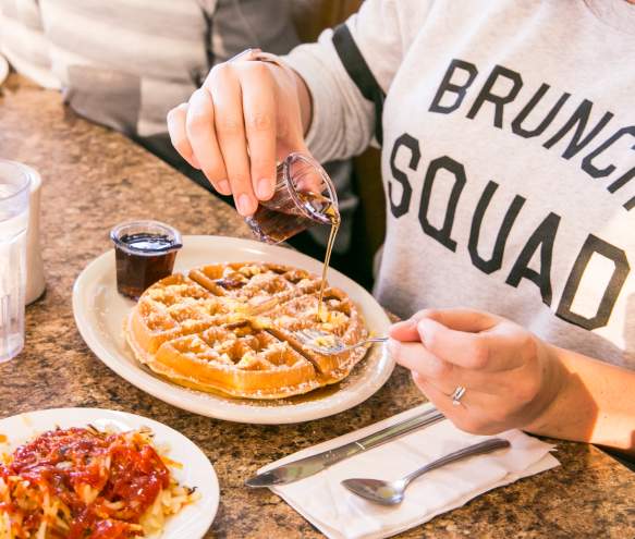 Person wearing a Brunch Squad shirt pouring maple syrup on a Belgian waffle at Cozy Table in Bloomington