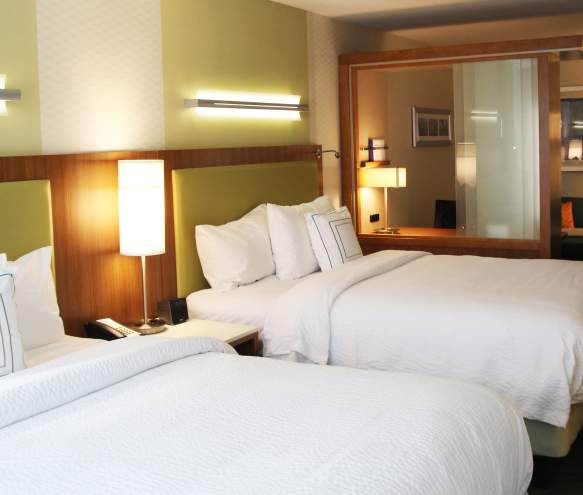 SpringHill Suites Hotel room interior with two neatly made beds, modern lighting, a bedside table, and a seating area with a sofa and lamp in the background.