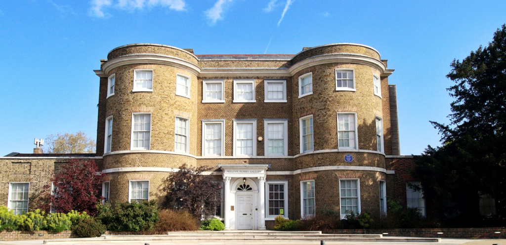Stately Georgian building with a curved façade, large windows, and a white door. Surrounded by green lawns and a clear blue sky overhead. William Morris Gallery in Lloyd Park, Walthamstow.