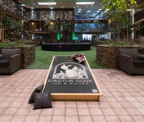 atrium of the cactus cove inn and suites. green putt putt area surrounding a black fountain and string lights above and a corn hole board with a cactus logo in the foreground