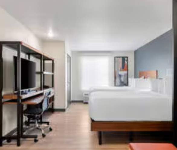 A modern hotel room featuring two queen beds with white linens, a work desk with a large TV, and a cozy sitting area separated by a patterned privacy screen. Light wood floors and clean lines give the space a bright, contemporary feel.