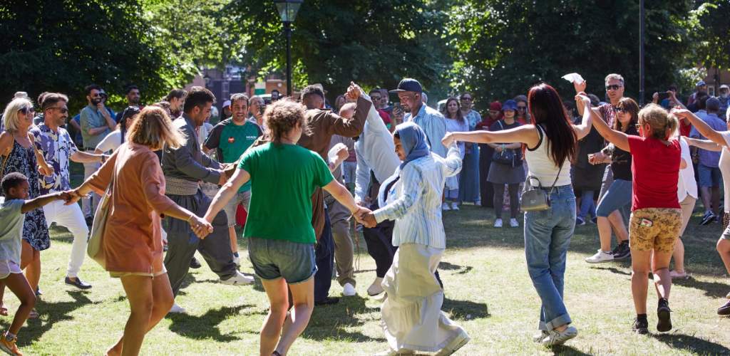 Group of people holding hands and dancing in a circle in Palmerston Park for Refugee Week