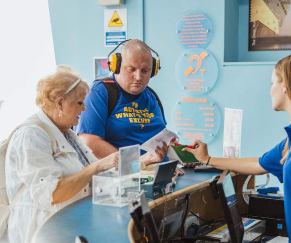 A staff member at a visitor attraction reception desk assisting two customers with tickets, highlighting accessible customer service and tourism support in a bright, welcoming environment.