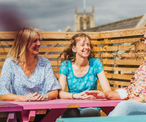 A man and woman  wearing a hearing aid relaxes group of friends at a colourful outdoor seating area on Humber Street in Hull. The scene showcases the vibrant, accessible social spaces of the Humber Street area
