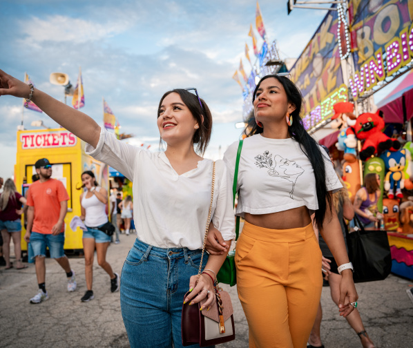 Two young women walk arm-in-arm through the Wisconsin State Fair, with one pointing excitedly ahead. Carnival rides, game booths, and crowds create a festive atmosphere in the background.