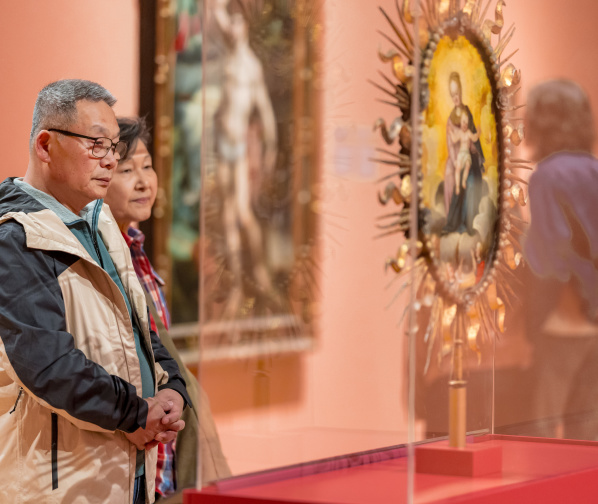 A middle-aged man wearing glasses and a tan-and-black jacket stands with his hands clasped, looking thoughtfully at an artwork in what appears to be an art museum or gallery. Behind him, a woman stands slightly out of focus, also observing the exhibit.

In the foreground on the right, there is a blurred religious-style artwork or icon displayed in a glass case, with a gold decorative frame. The overall scene has warm lighting and a calm, contemplative mood.
