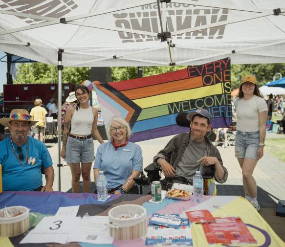 Five people smile under a tent at a community Pride event, seated at an information table with a rainbow ‘Everyone is welcome here’ flag displayed behind them.