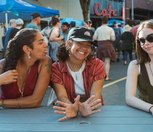 Three women sit at a picnic table at a busy outdoor summer market. One playfully sticks out her tongue toward her friend while another smiles and gestures toward the camera.