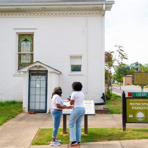 Two women are standing and holding a phone in front of the Kentucky Black Trailblazers plaque for The First Black Baptist Church located in downtown Elizabethtown.