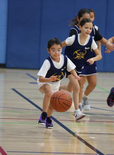 Young girl dribbles basketball indoors with her team and opponent