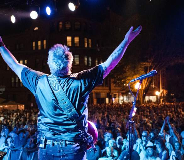 guitar player on stage with hands up in front of huge crowd at night