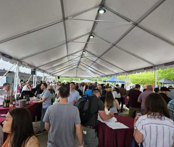 A crowd of people gather under a tent at the Wine & Food Festival to savor their food samples.