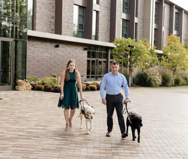 Two people walk on an outdoor stone pathway with service dogs. A building and trees are in the background.