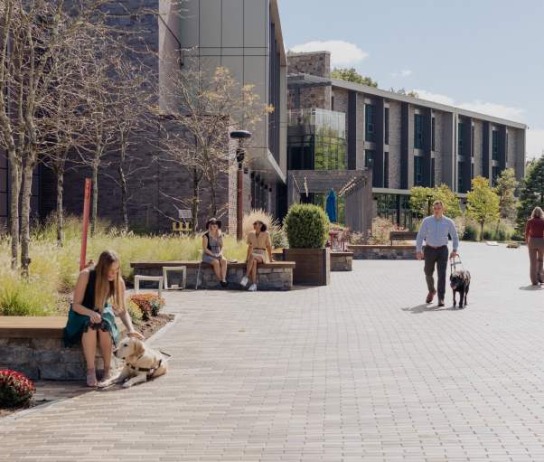 A woman is seated outside in summer, smiling and petting a service dog. Another man is walking toward the woman with the service dog. Two people are walking away toward a building in the distance.