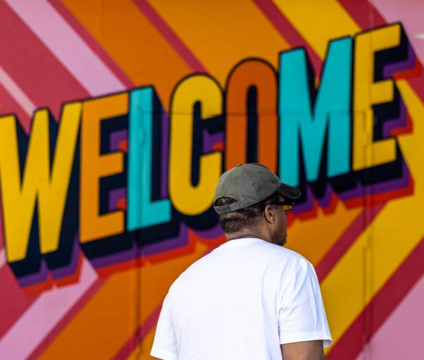 A man stands in front of a multi-colored welcome sign.