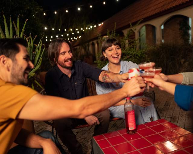 Group of five men and women sit at the Austin Motel courtyard under string lights at night and cheers with cocktails and beers.