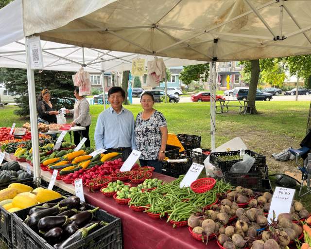 Vendors stand behind a table filled with fresh vegetables—eggplant, squash, beans, radishes and more—at a neighborhood farmers market, with tents, trees and nearby homes in the background.