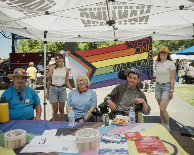 Five people smile under a tent at a community Pride event, seated at an information table with a rainbow ‘Everyone is welcome here’ flag displayed behind them.