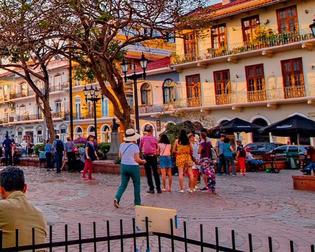 The image shows a vibrant street scene in a historic city. The street is lined with colorful buildings, some with balconies and ornate details. There are people walking around, enjoying the evening. In the background, there is a church with a blue facade and a bell tower.