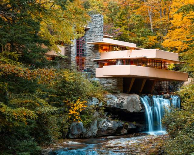 Iconic architectural building Fallingwater sits atop a waterfall with fall foliage surrounding.