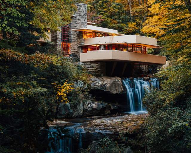 A modern, tan home , known as Frank Lloyd Wright's Fallingwater, situated on top of a waterfall with fall foliage-filled trees surrounding it