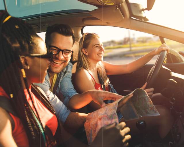 Three people in a car. Two women sit in the front seat with a man poking his head up front from the backseat with a map