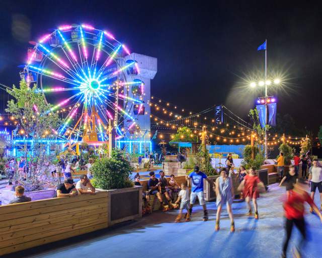 People rollerblading on a rink at night with string lights overhead and a glowing ferris wheel in the background.