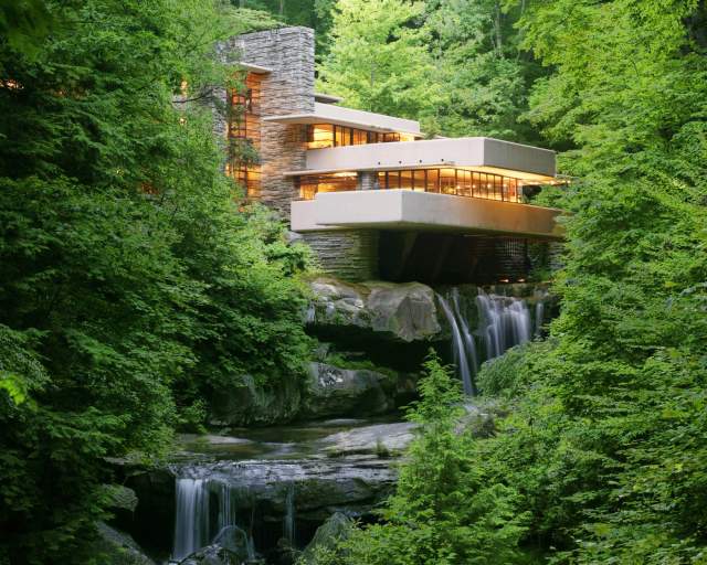 A modern home sitting on top of a waterfall with green trees surrounding it