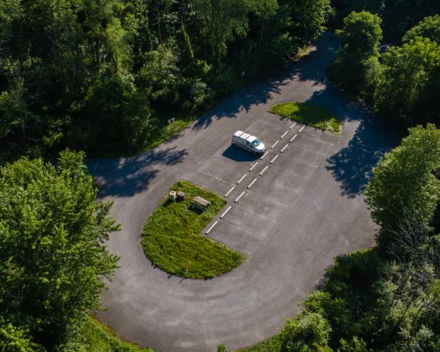 Aerial view of a oval-shaped parking lot. A lone white van is parked. Trees surround the lot.