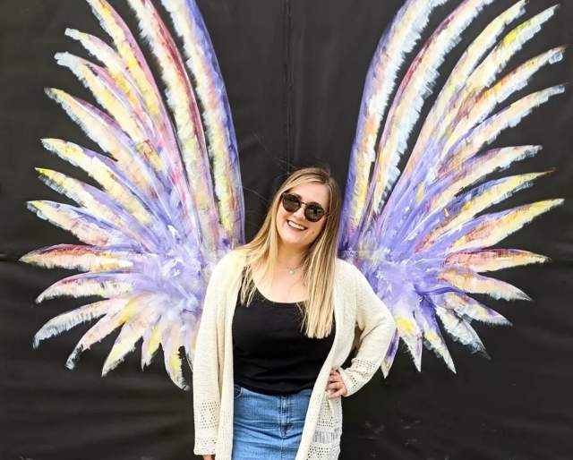 A smiling woman in sunglasses poses in front of a colorful painted mural of angel wings on a black backdrop in Fayetteville, NC. The wings are lavender with accents of yellow, pink, and white, creating a vibrant, photo-worthy moment.