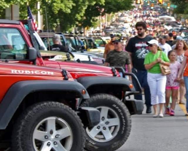 Red jeeps lined up in a row with a crowd of people in the street walking past.