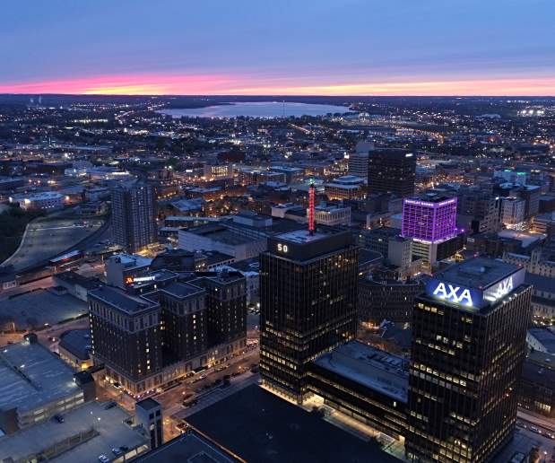 Syracuse city skyline at dusk with purple sky in the background, AXA towers and Barclay Damon building in the foreground, lit up purple.  City lights illuminate the photograph