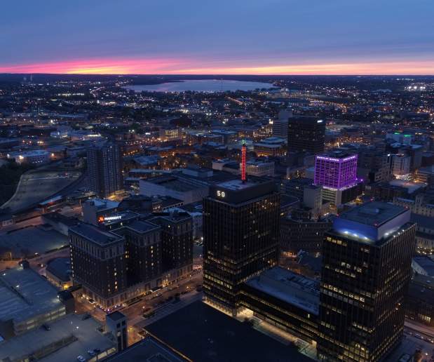 Syracuse city skyline at dusk with purple sky in the background, AXA towers and Barclay Damon building in the foreground, lit up purple.  City lights illuminate the photograph