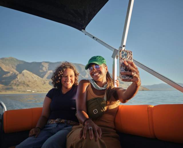 Two women on a pontoon boat taking a selfie with the great salt lake and antelope island in the background