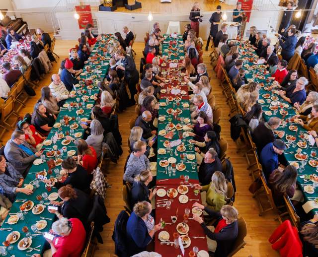 Aerial view of long tables set with red and green table cloths with people sitting at them, eating, and talking.