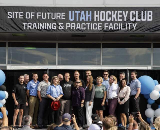 A banner hanging reading 'site of future Utah Hockey Club Training and Practice Favility. a group of executives stainging underneith with a crowd.