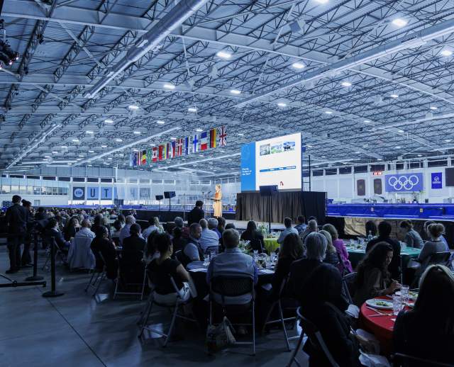 Speaker on stage at the Utah Olympic Oval, people sitting at round tables in the foreground, UT Hockey Club and Olympic logos in the background